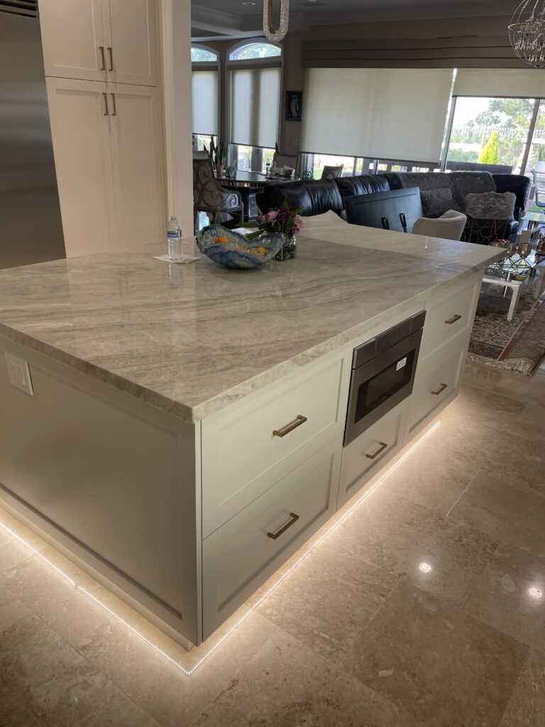 White and cream marble top on the kitchen island with complimnetary beige drawers and clean stainless steel mini oven, microwave in the middle of the kitchen island. Beautiful LED lights under the island countertops that gives the kitchen a modern and classy look. With marble flooring.