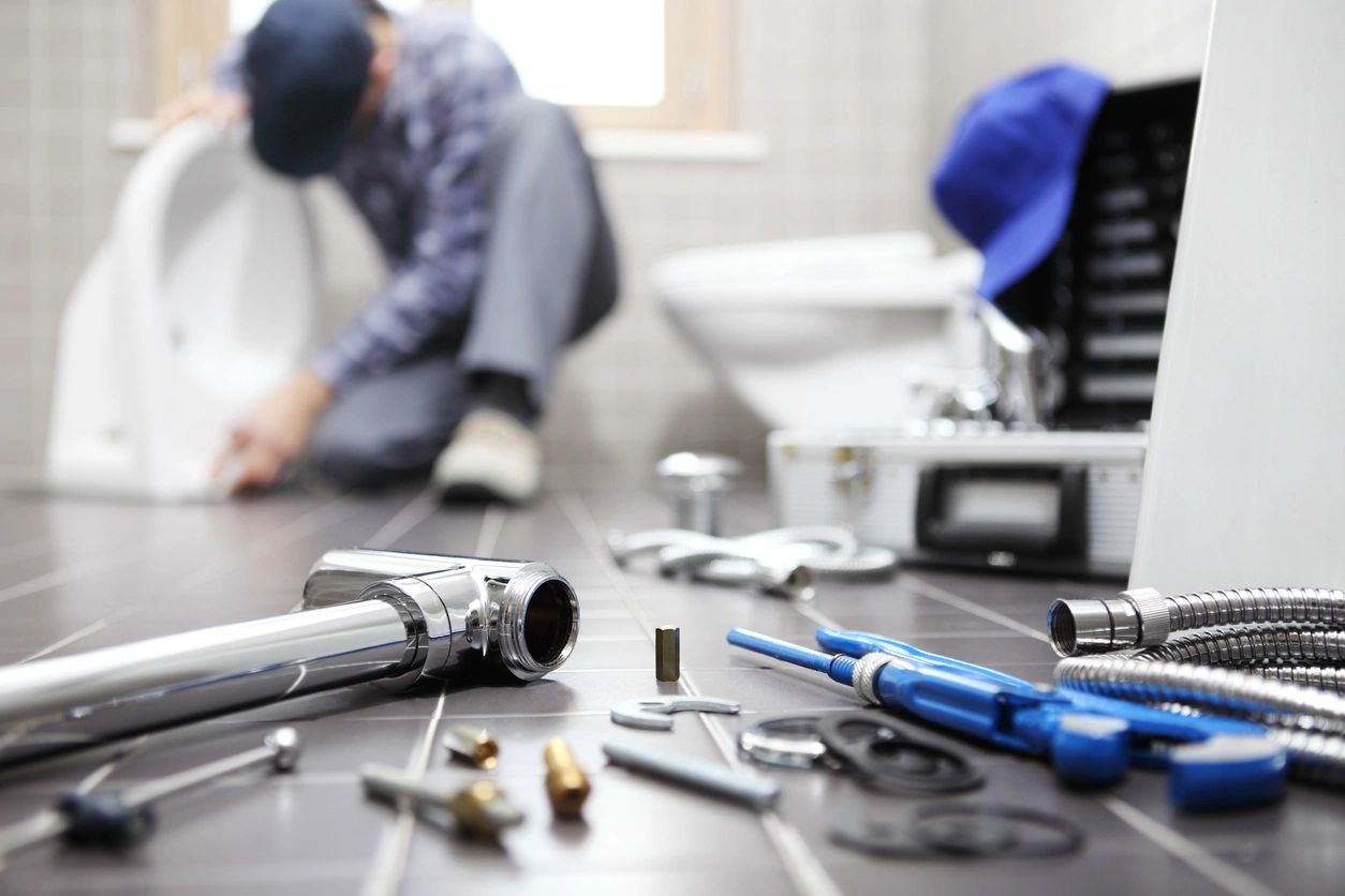 black floor with the focus on the plumbing tools at the front and blurred background with a white toilet and blurry plumber next to the toilet working.